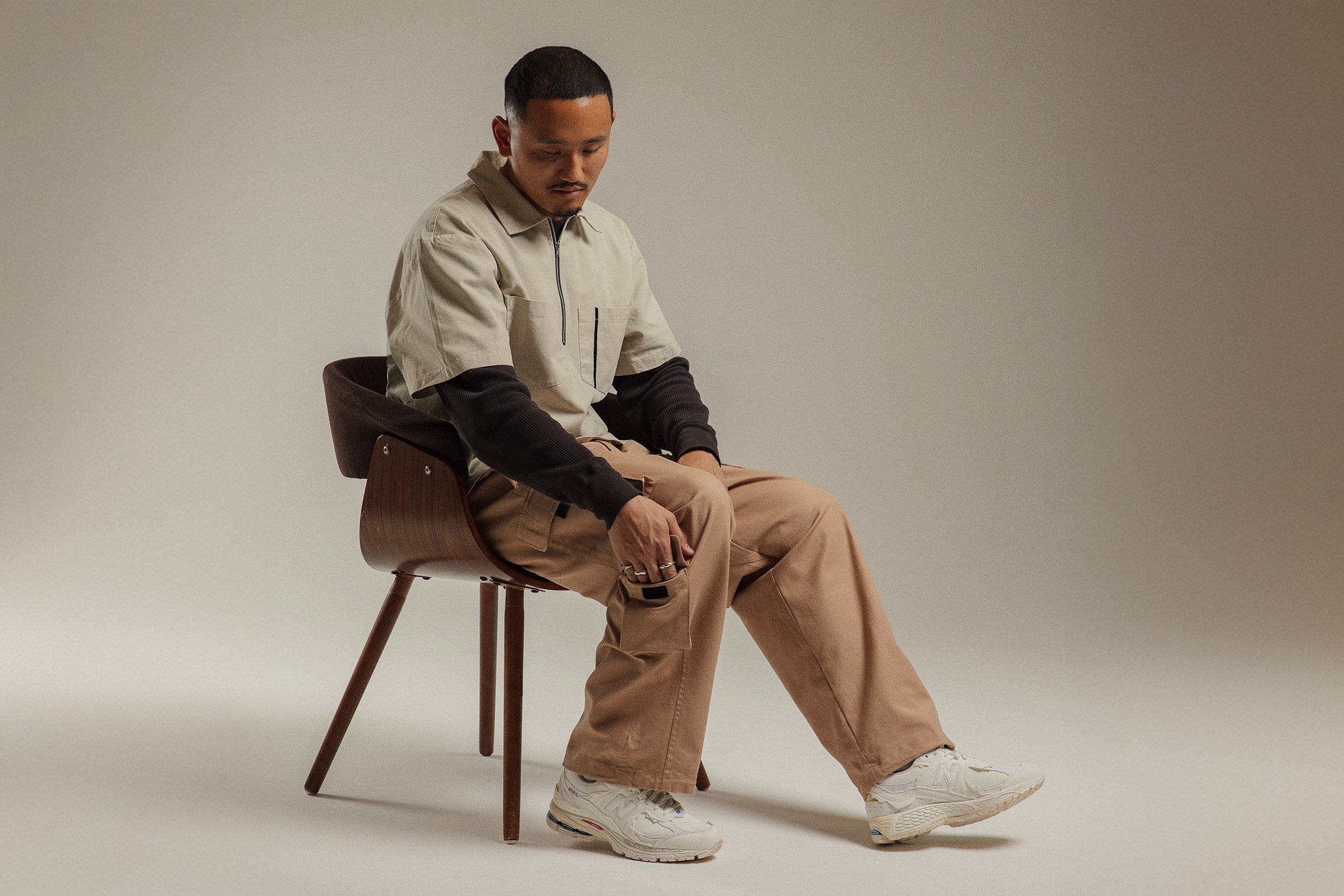 Man sitting on a chair wearing a light jacket and beige pants against a plain background