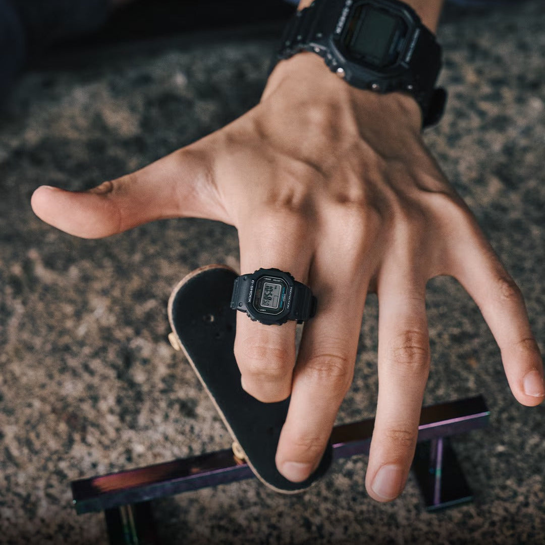 Hand wearing a G-Shock watch with a skateboard on a textured surface