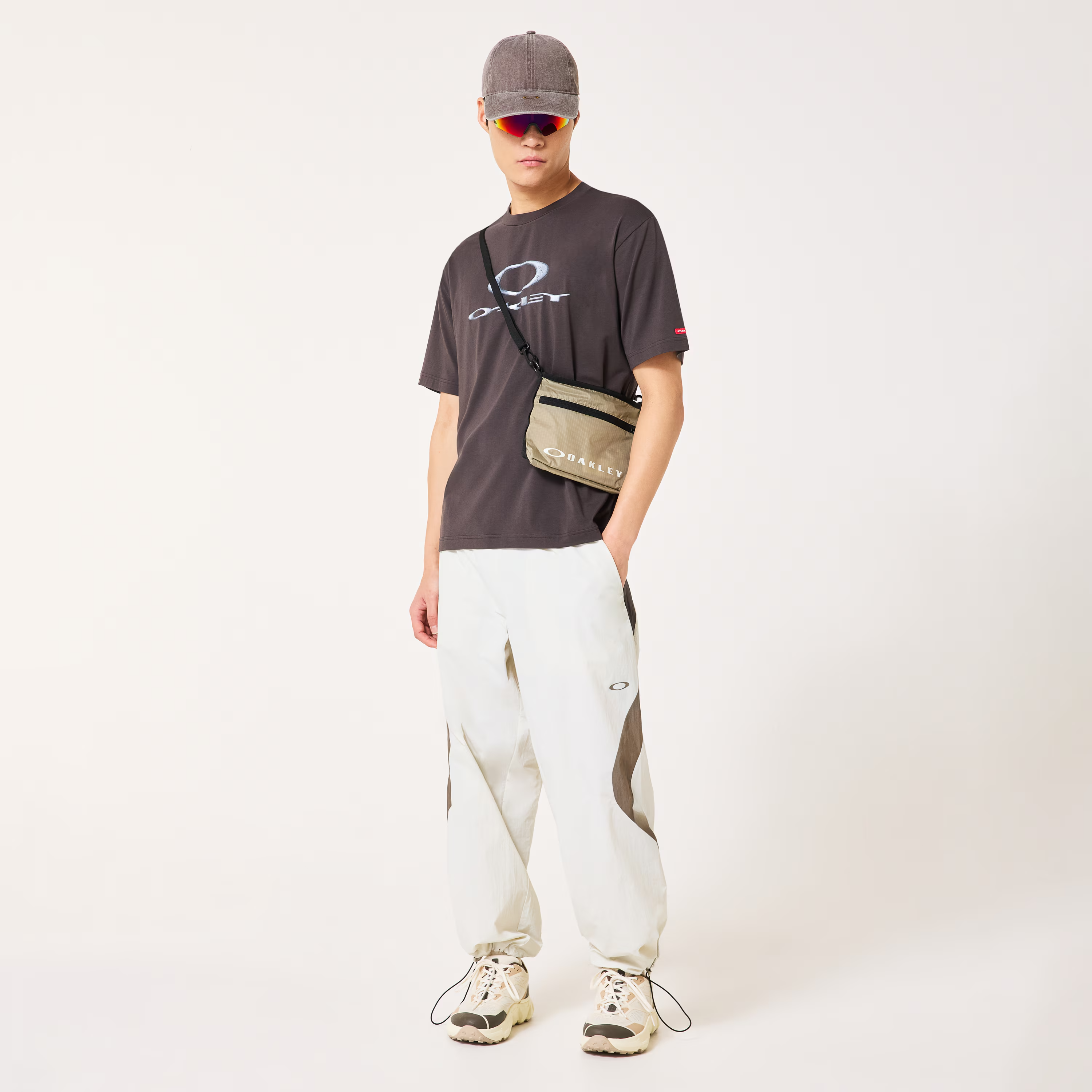 A model wearing a dark brown Oakley t-shirt, white cargo pants with brown side panels, a brown cap, and sunglasses, carrying a brown Oakley crossbody bag.